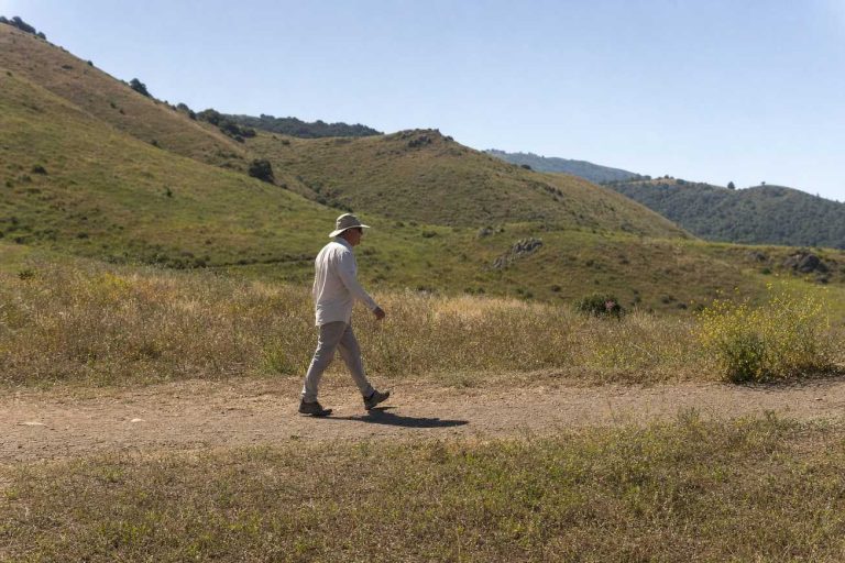 An older hiker in a long-sleeve shirt and wide-brimmed hat walking an open, sunlit trail