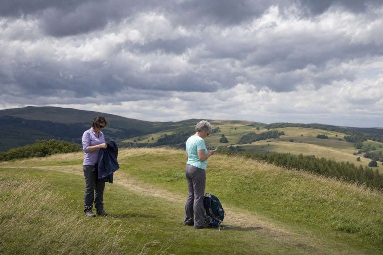 Two hikers pause on an open trail as dark clouds build over the hills behind them