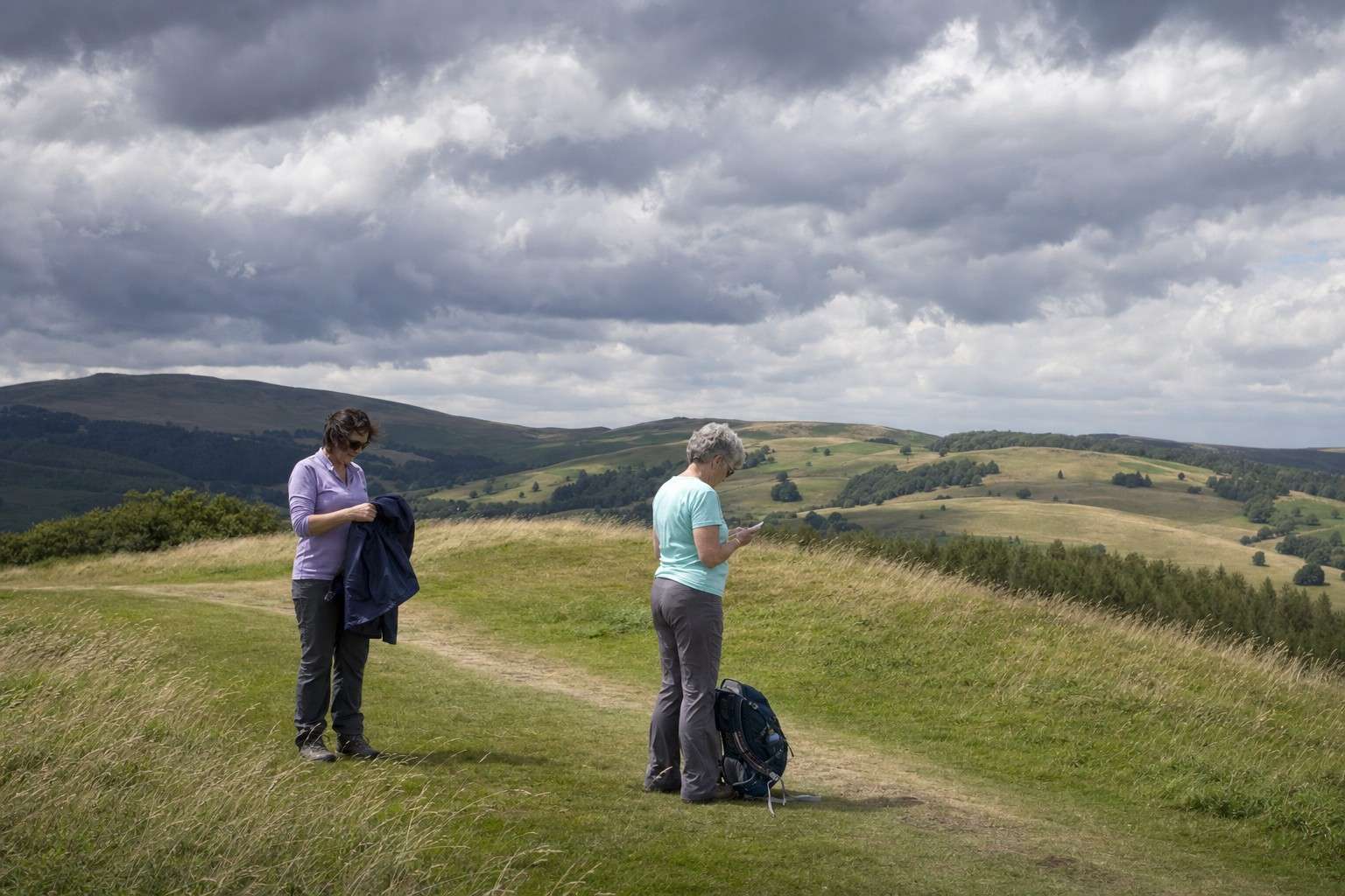 Two hikers pause on an open trail as dark clouds build over the hills behind them