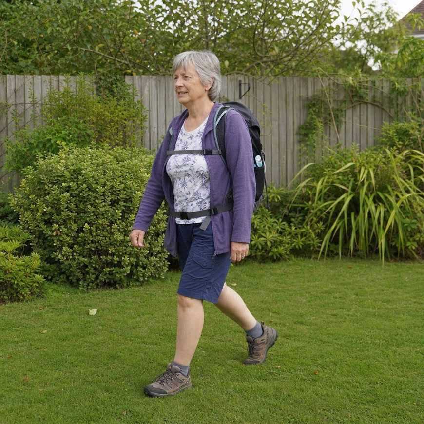 An older woman walking across her garden wearing a small loaded daypack, standing tall