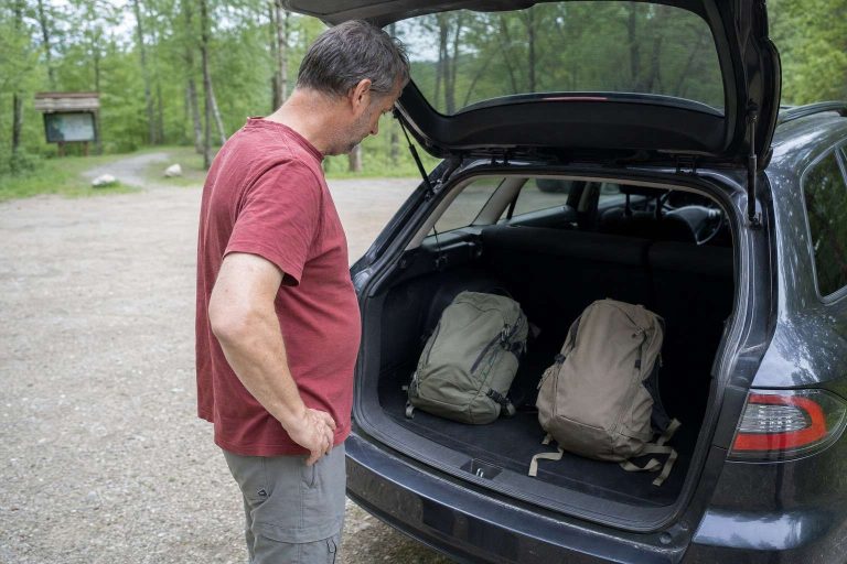 A man over 50 deciding between two small daypacks at the open tailgate of his car