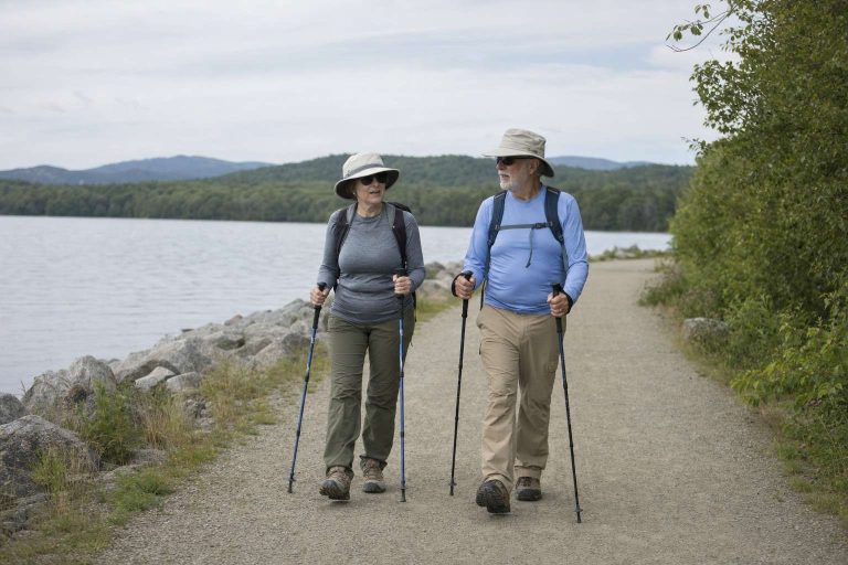 Two older hikers walking toward the camera on a long lakeside trail